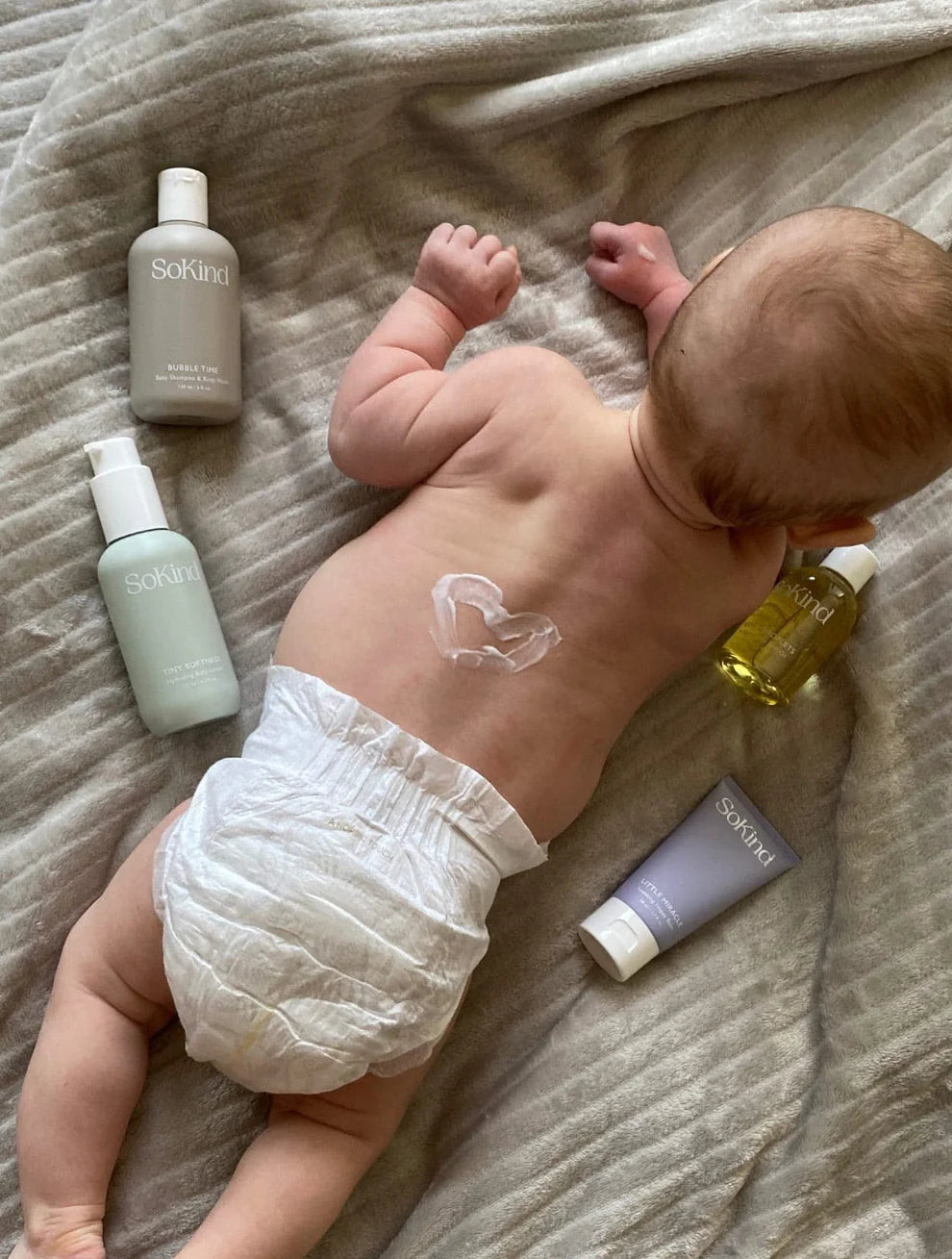 Een baby met de vier verzorgingsproducten uit de SoKind Dear Baby Skin Care Kit rond zich en een hartje op zijn rug getekend in verzorgingscrème.