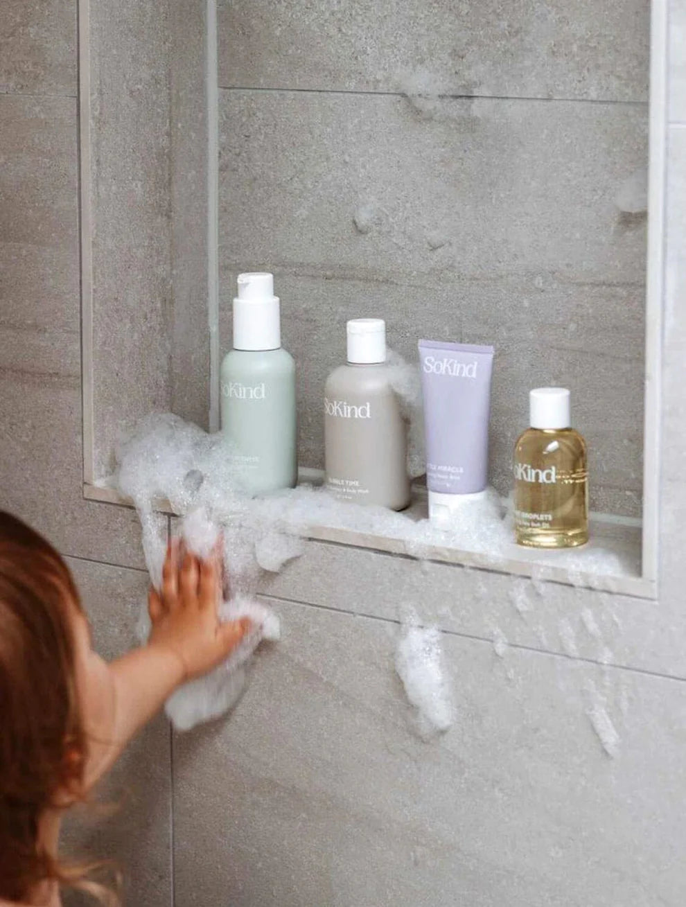 De verzorgingsproducten uit de SoKind baby verzorgingsset uitgestald in de douche, waarnaar een kindje in de douche grijpt.