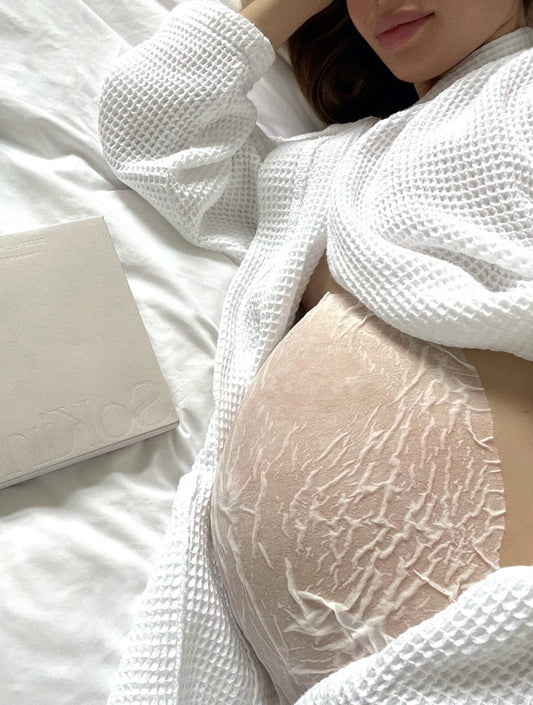 Hoogzwangere vrouw ligt op bed met het Blissful Moment Belly Sheet Masker van SoKind op haar buik.